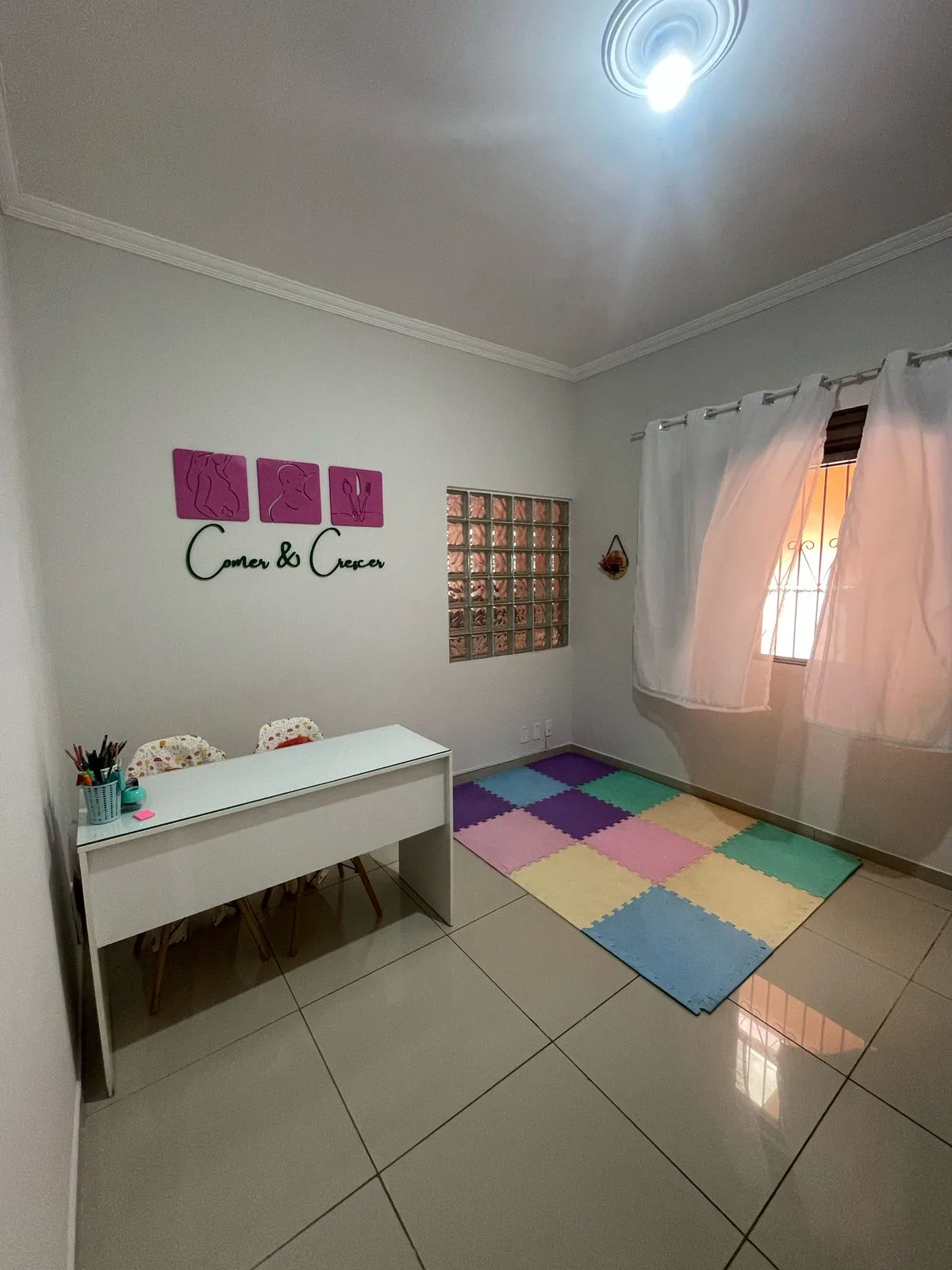 Consultório da Comer e Crescer: mesa branca, tapete colorido e, na parede, os três quadros magenta do logo com o nome Comer e Crescer